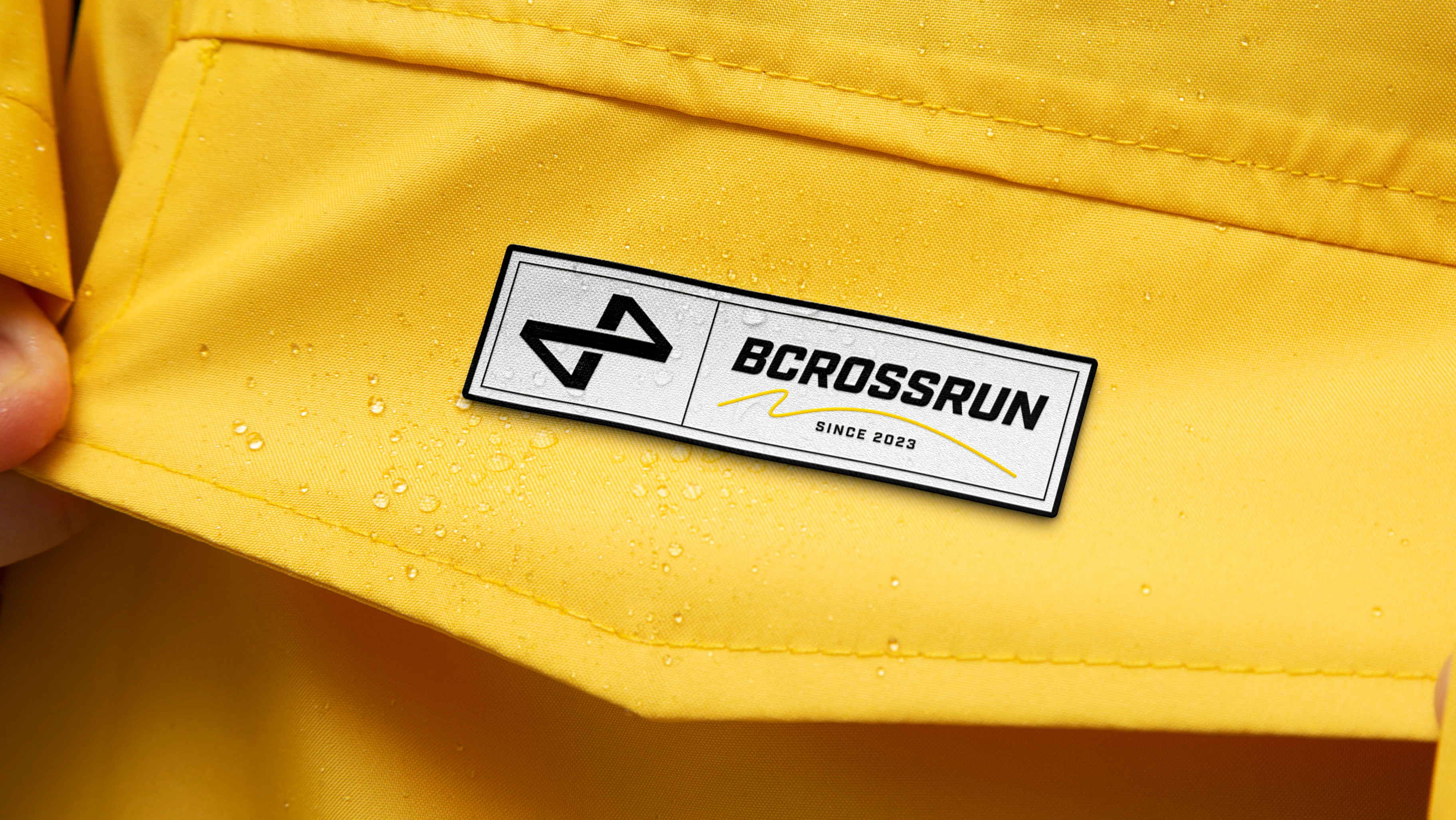 Bcrossrun branded label on yellow technical jacket — black-and-white woven badge reading BCROSSRUN Since 2023 with water droplets