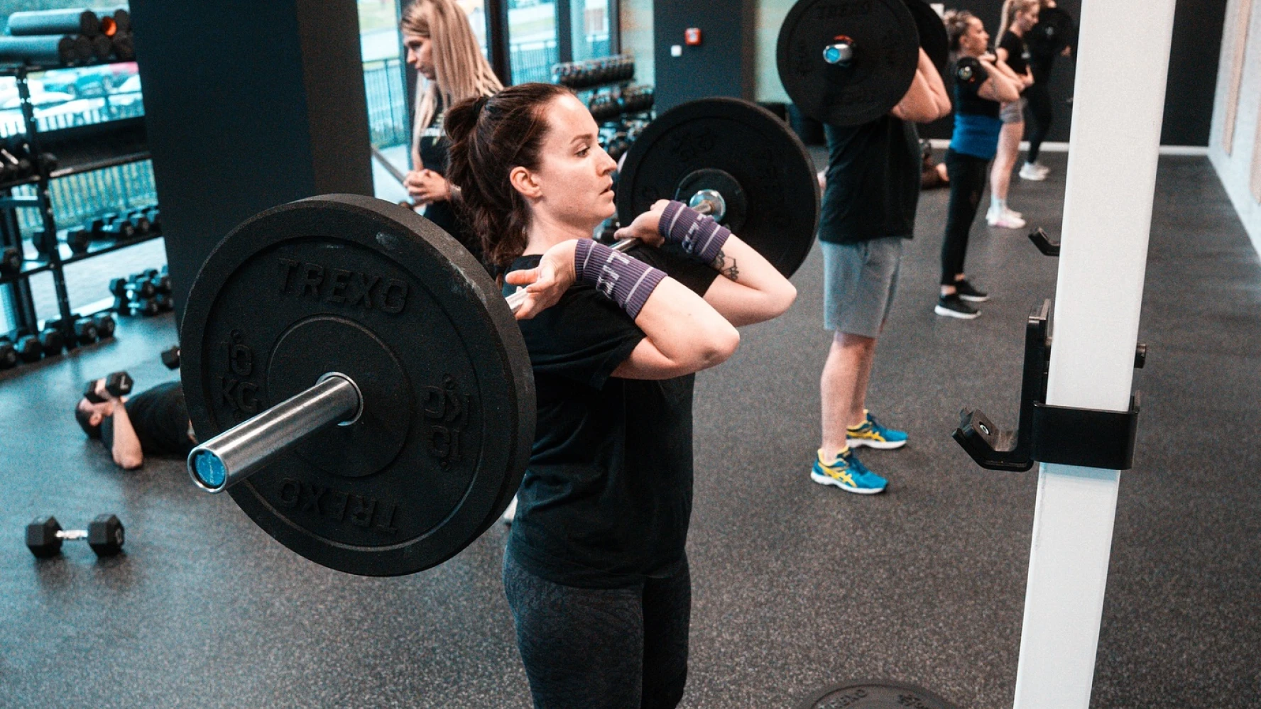 Fitness Central Park gym photography — female athlete front-squatting with TREXO barbell amid an open class on the training floor