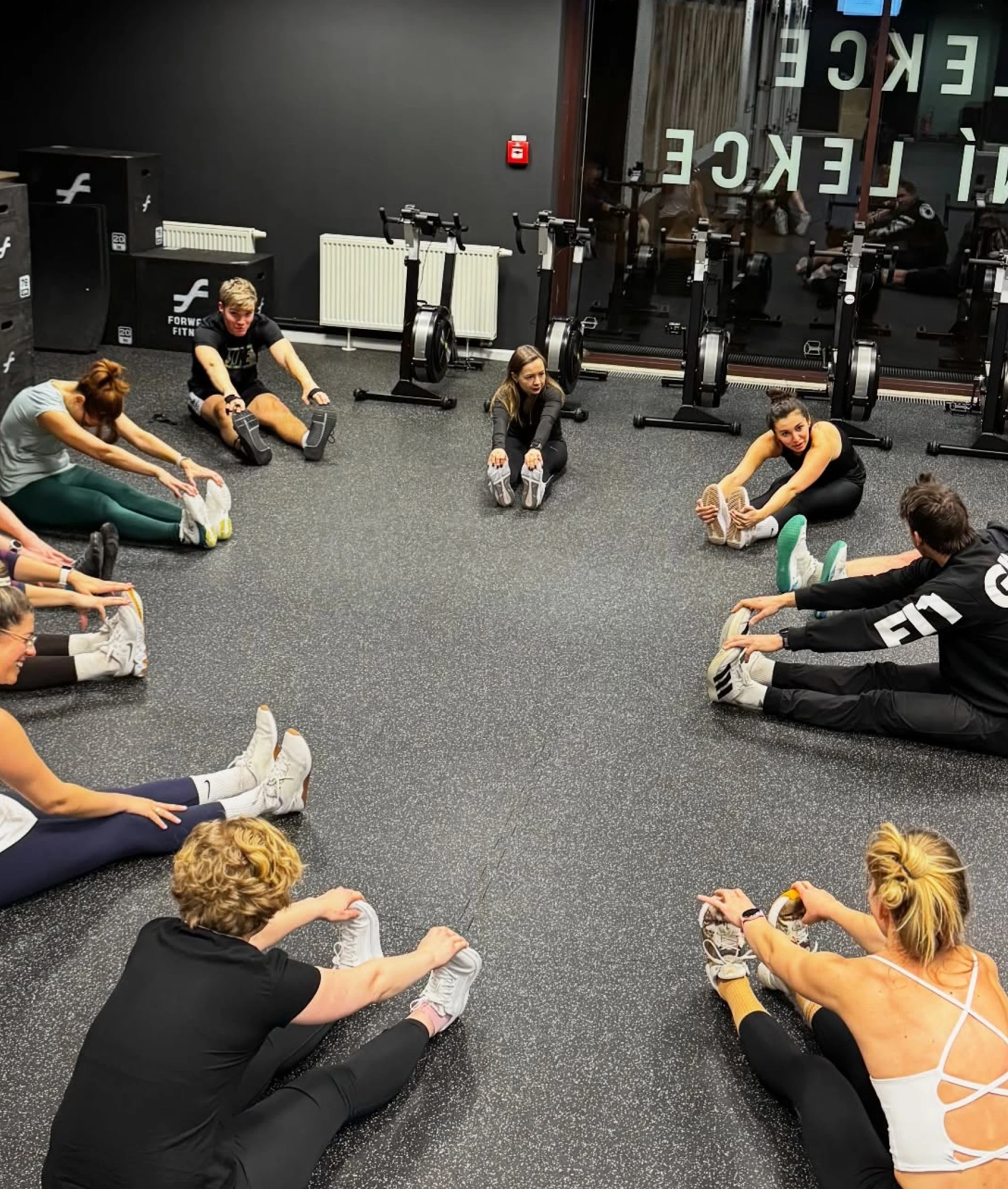 Fitness Central Park group session photo — circle of athletes stretching on the rubber floor with rowers and LEKCE wall graphics in the background