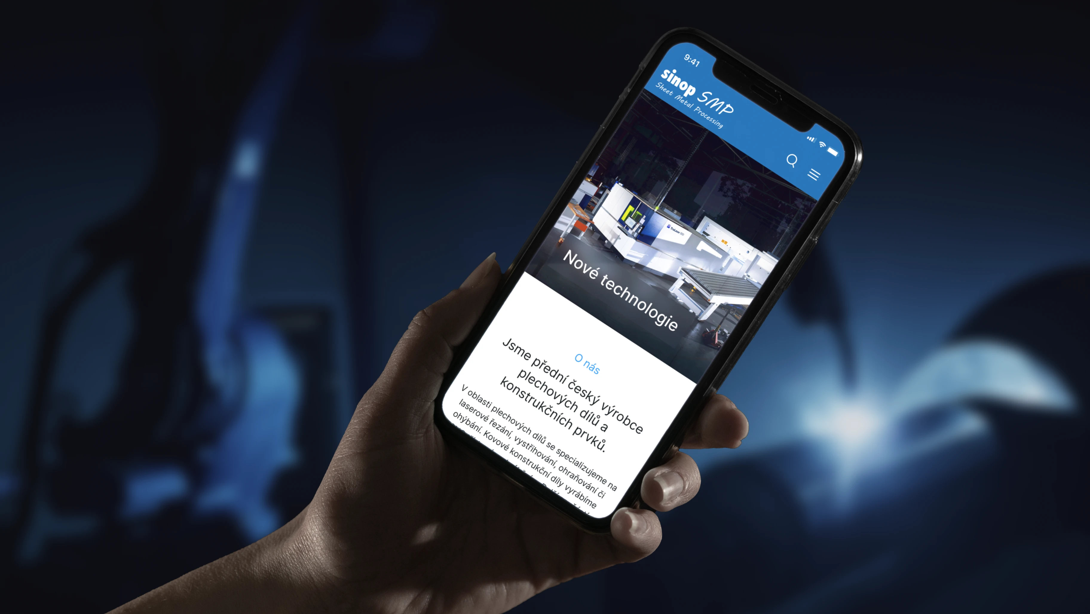 Sinop s.m.p. mobile experience — hand holding iPhone showing the Sinop SMP Nové technologie homepage against a deep blue out-of-focus factory backdrop