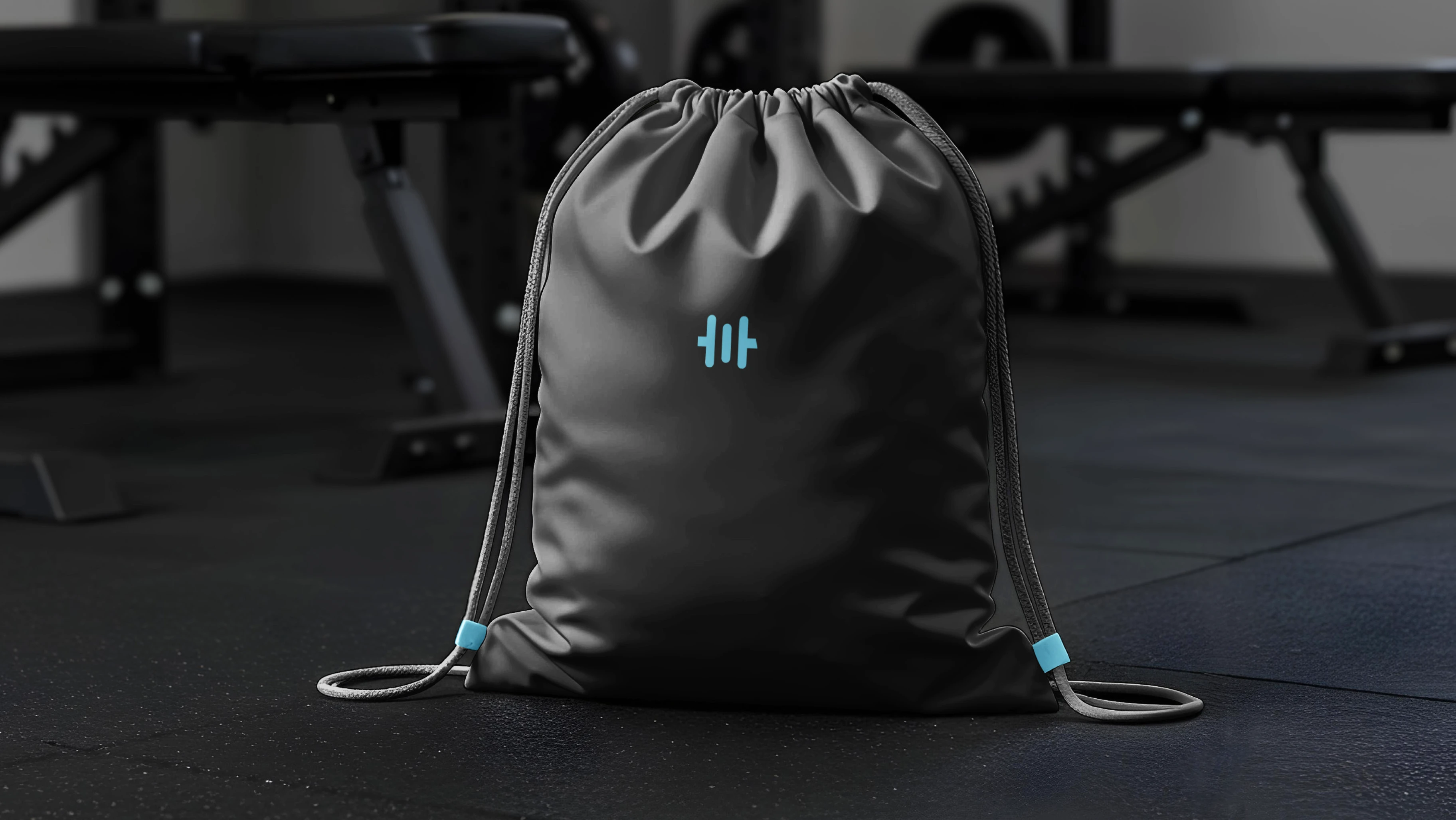 Wodcomp drawstring competition bag — black sports bag with Viking-blue dumbbell-grip logo and matching tab accents standing on rubber gym flooring